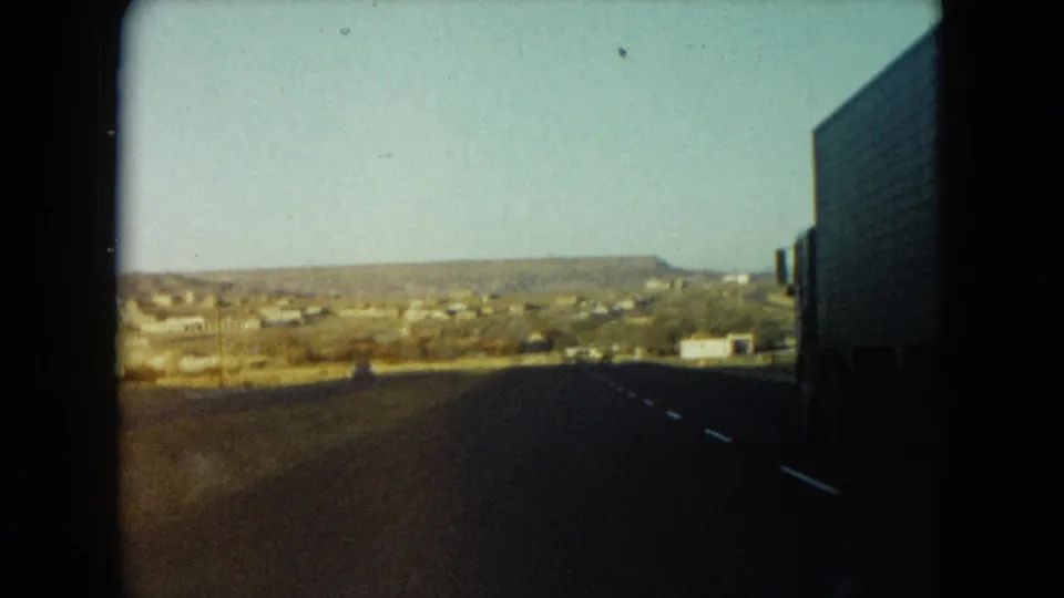 Moving 1960 Nevada highway view toward distant low buildings, with a large truck passing at the right edge.