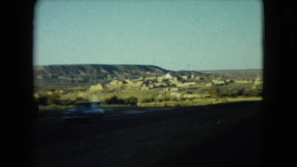 1960 Nevada 8mm roadside view with distant buildings, hills, utility poles, and a blurred car passing in front.
