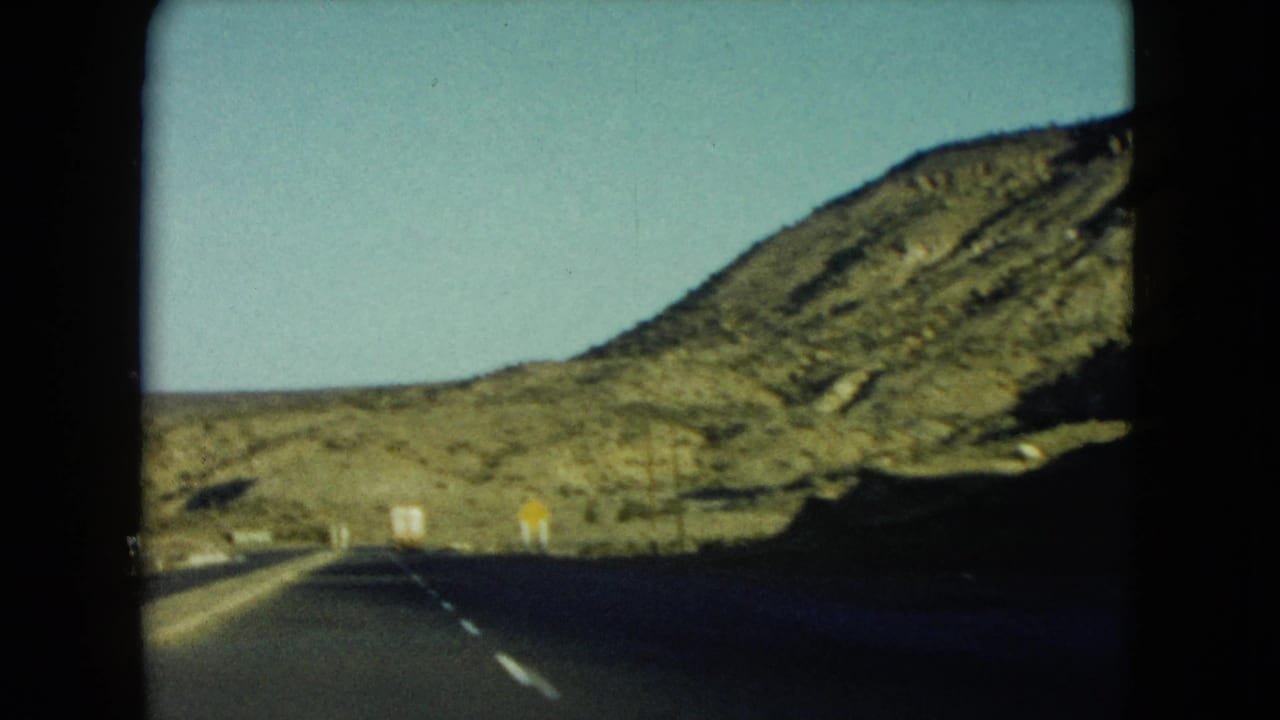 1960 Nevada 8mm view down a two-lane highway toward rocky desert hills and small roadside signs.