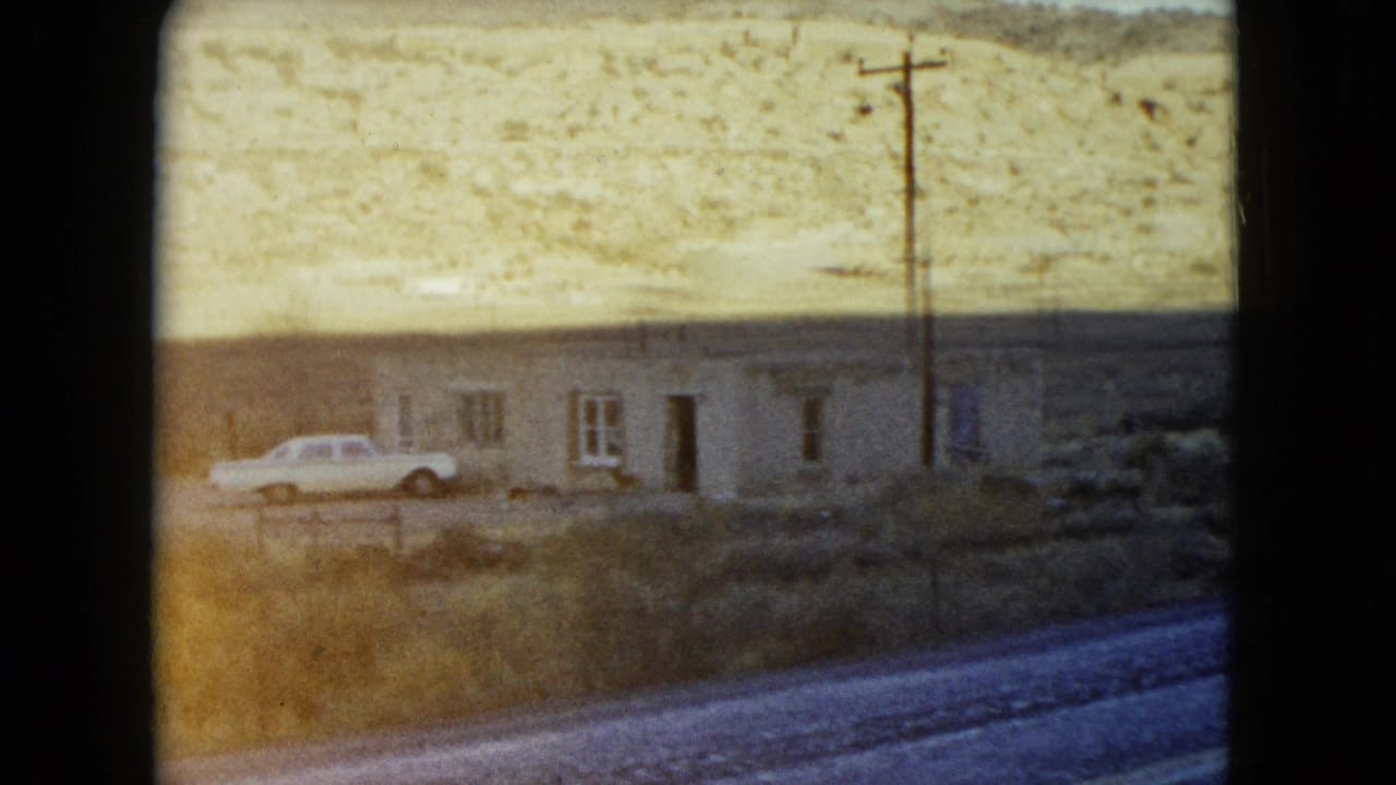 1960 Nevada roadside scene with a low building, parked light-colored car, utility pole, and dry hillside beyond.