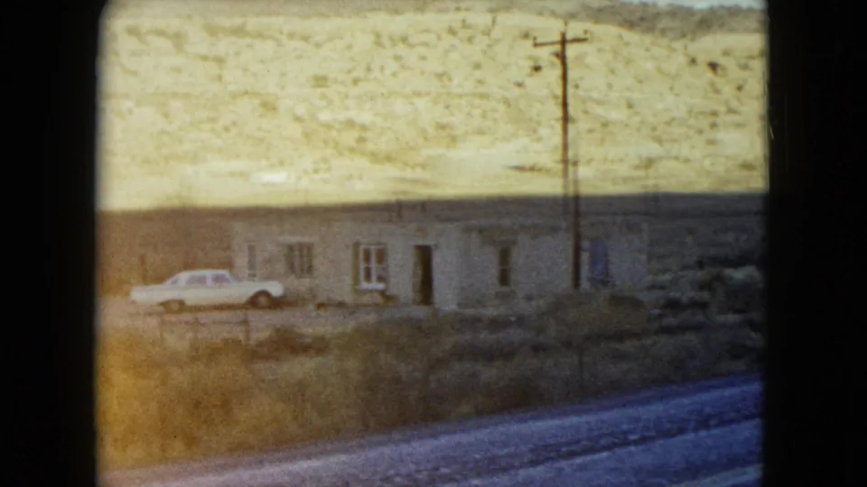 1960 Nevada roadside scene with a low building, parked light-colored car, utility pole, and dry hillside beyond.