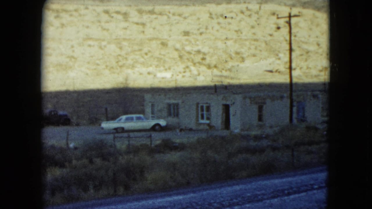 White car parked beside a low stony house below a pale desert hillside.