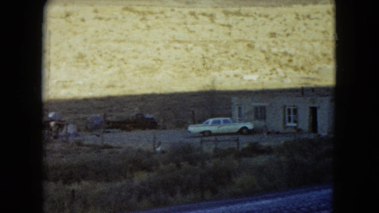 1960 Nevada frame with a white parked car, low building, sparse scrub, and muted desert hills in the background.