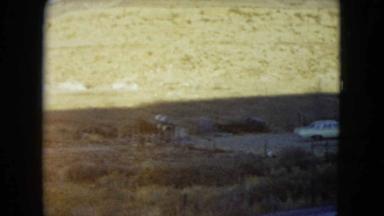 Soft 1960 Nevada 8mm frame of a remote roadside building area, scrubland, hillside, and a white car partly at the right edge.