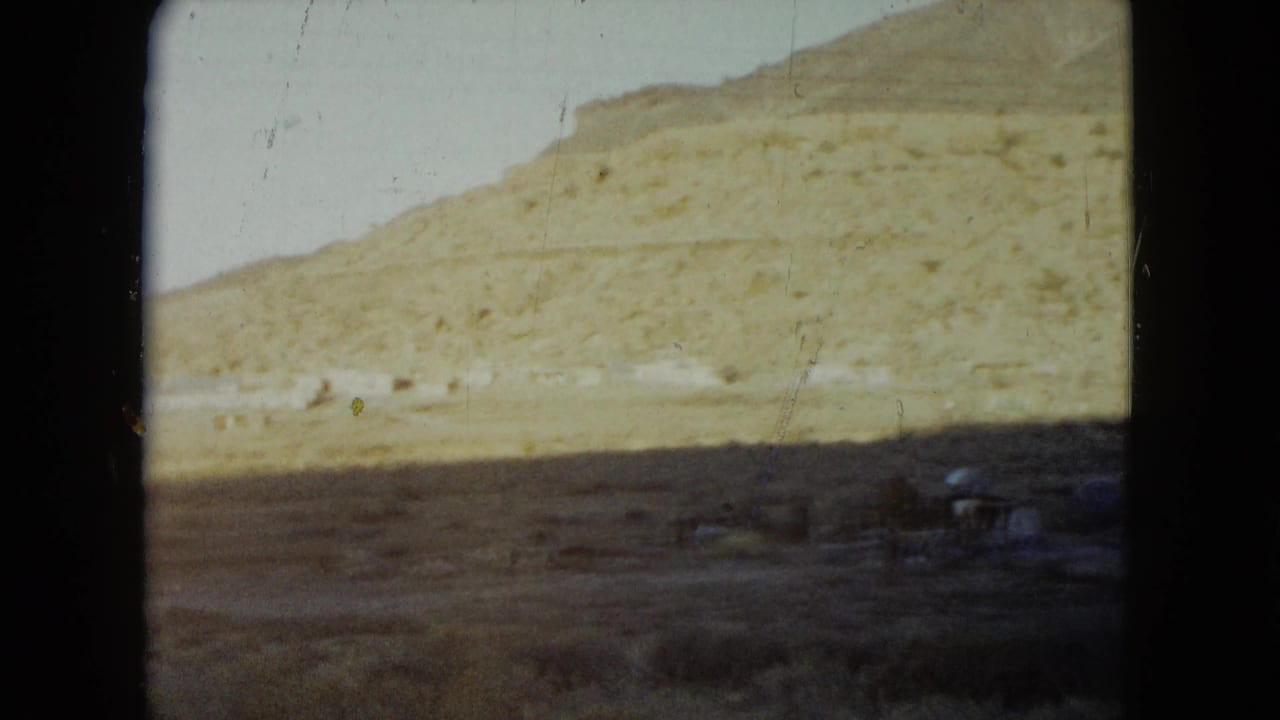 1960 Nevada 8mm frame looking across a dry hillside and sparse foreground with small roadside structures near the bottom of the frame.