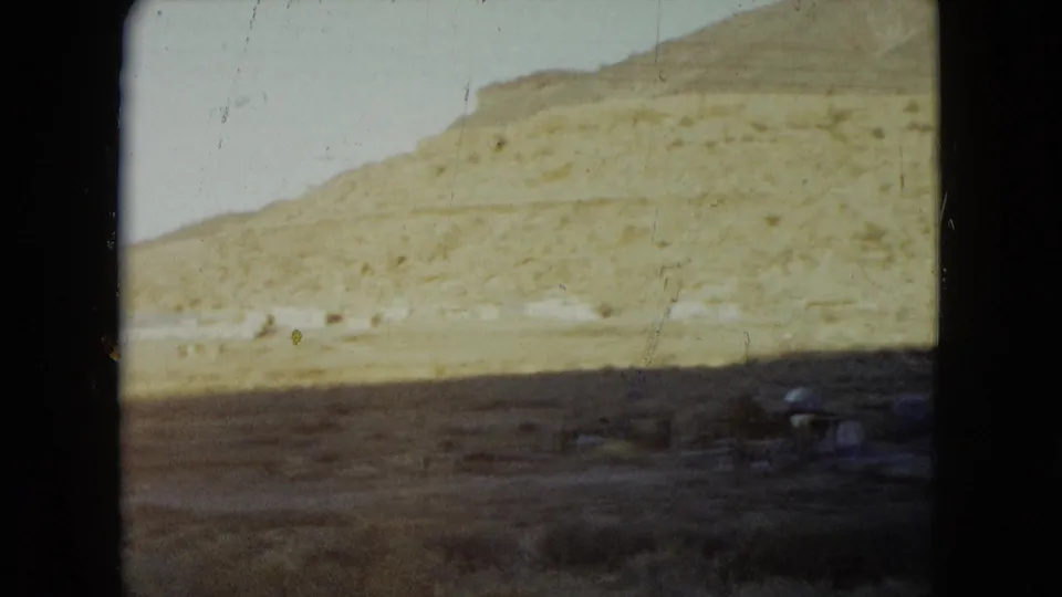 1960 Nevada 8mm frame looking across a dry hillside and sparse foreground with small roadside structures near the bottom of the frame.