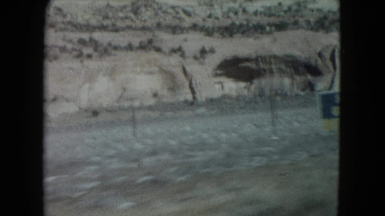 Moving-car view in Nevada showing blurred pavement, pale cliff layers, scrub, and a roadside structure or sign edge.