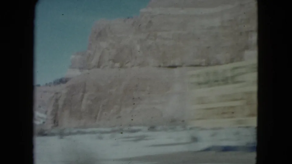 1960 Nevada road-view frame with layered sandstone cliffs, open sky, and motion-blurred desert terrain below.