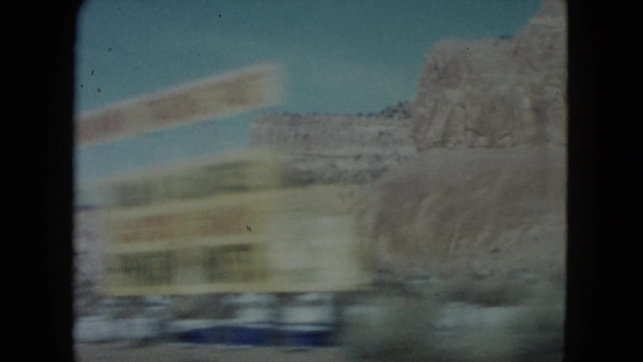 1960 Nevada 8mm frame with a yellow roadside sign streaking past the camera and sandstone terrain behind it.
