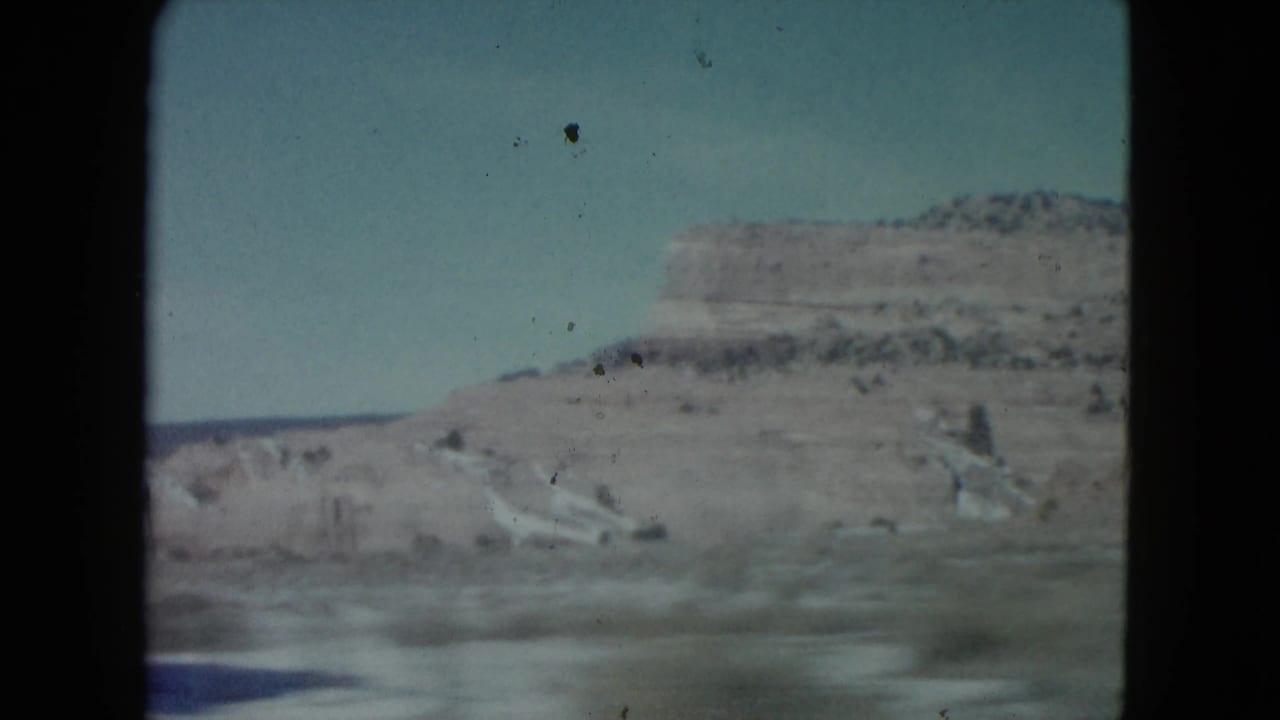 1960 Nevada 8mm frame showing a distant desert ridge, pale roadside terrain, sparse vegetation, and blue sky from a moving car.