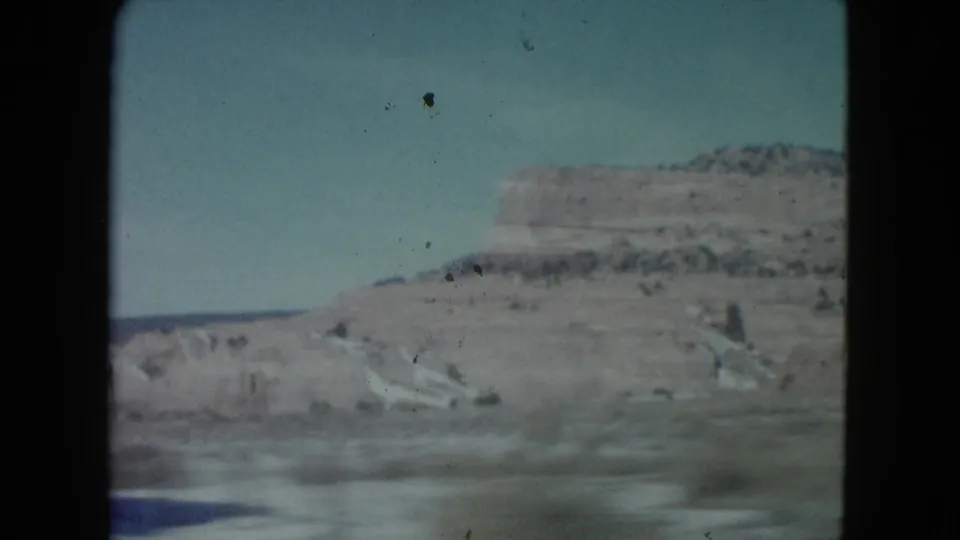 1960 Nevada 8mm frame showing a distant desert ridge, pale roadside terrain, sparse vegetation, and blue sky from a moving car.