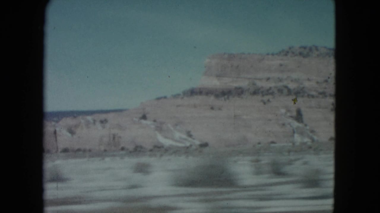 1960 Nevada desert road view with pale sky, layered mesa, and blurred foreground from a moving vehicle.