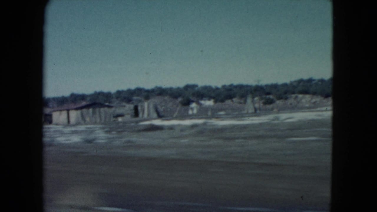 1960 Nevada roadside scene showing a small low building, open ground, and distant tree line or brush.