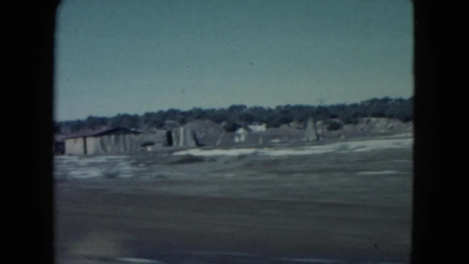 1960 Nevada roadside scene showing a small low building, open ground, and distant tree line or brush.