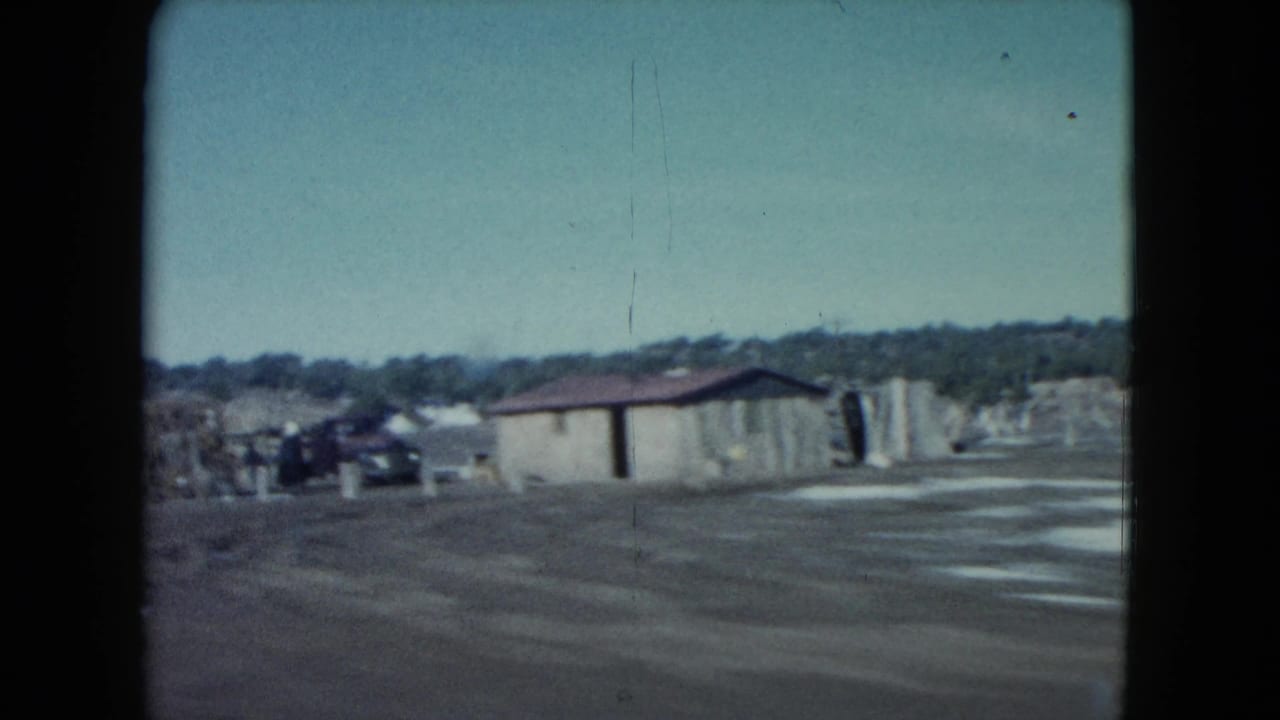 Nevada home-movie road view passing a small roadside building with distant structures under a pale sky.