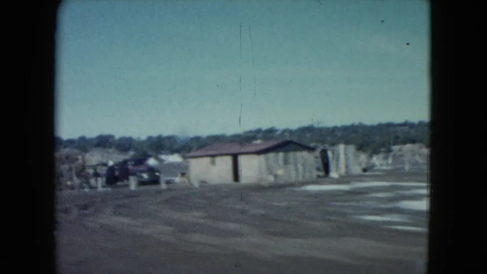 Nevada home-movie road view passing a small roadside building with distant structures under a pale sky.