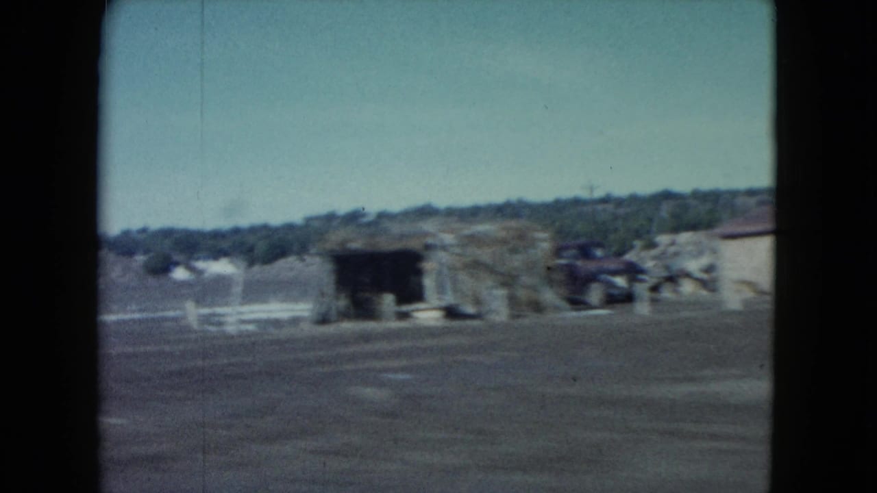 1960 Nevada 8mm frame showing a remote roadside scene with low adobe-like structures, dry ground, sparse vegetation, and distant hills.