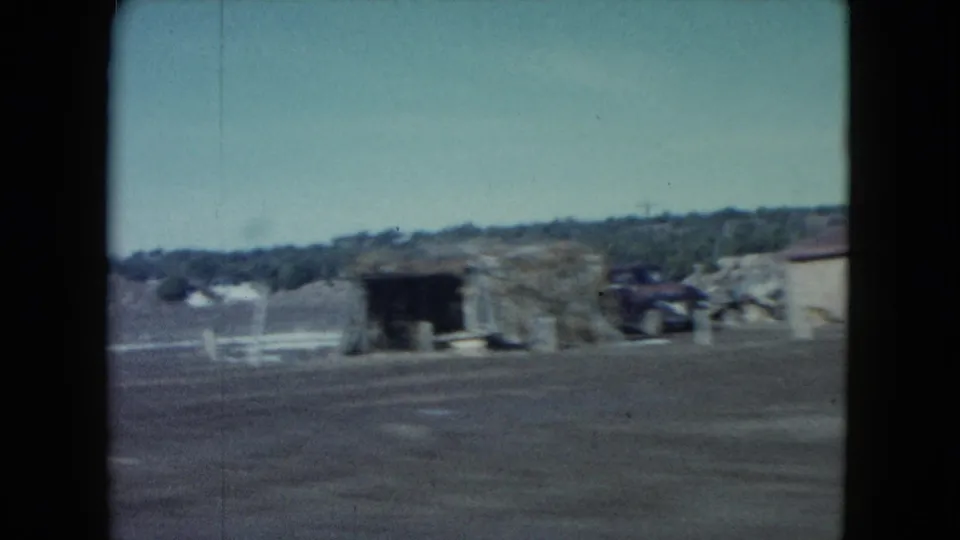 1960 Nevada 8mm frame showing a remote roadside scene with low adobe-like structures, dry ground, sparse vegetation, and distant hills.