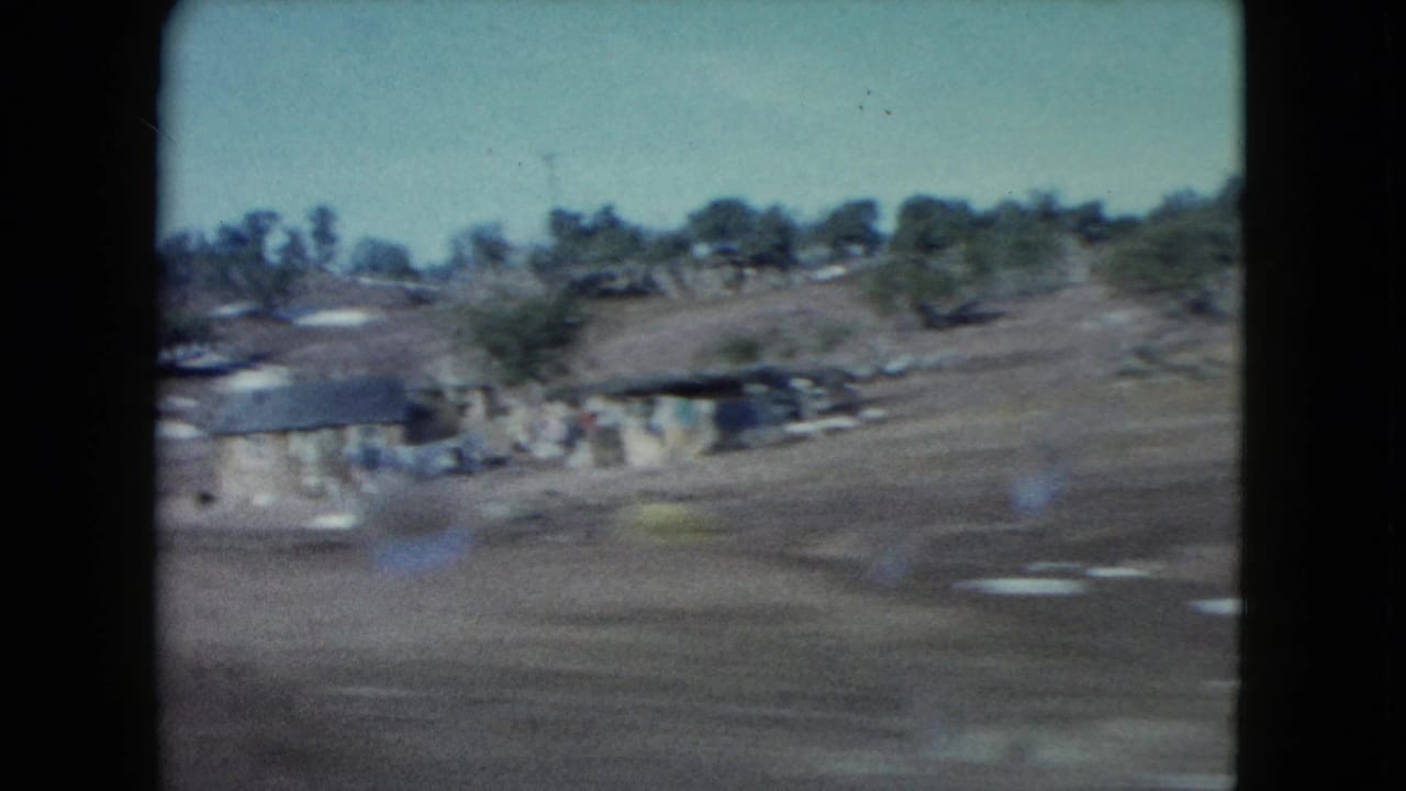 1960 Nevada home-movie frame with low rural buildings, open desert ground, scattered trees, and a scrubby hillside in the distance.
