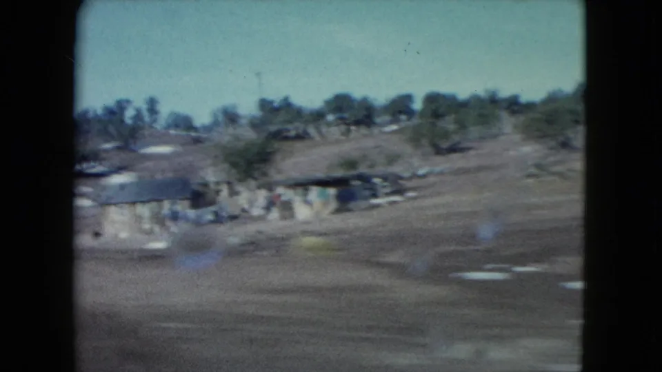 1960 Nevada home-movie frame with low rural buildings, open desert ground, scattered trees, and a scrubby hillside in the distance.