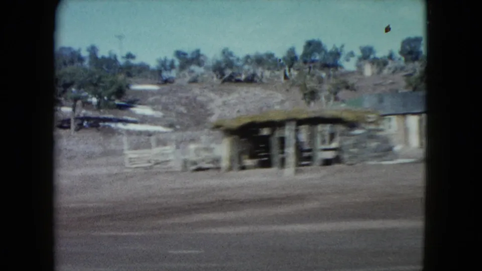 1960 Nevada 8mm view of a blurred low roadside structure, fence line, dry terrain, sparse trees, and low desert hill.