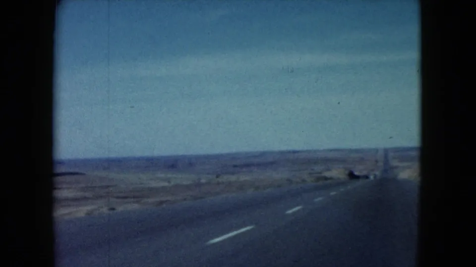 1960 Nevada 8mm frame showing an open paved road through flat desert, with utility poles, distant horizon, and wide blue sky.
