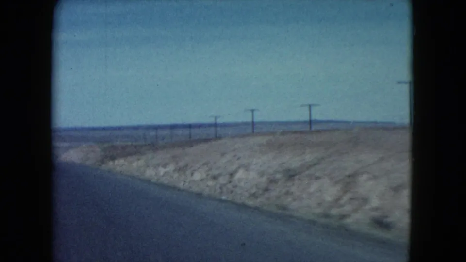 A moving-car view along the edge of a desert highway, showing pale soil banks and roadside utility lines.