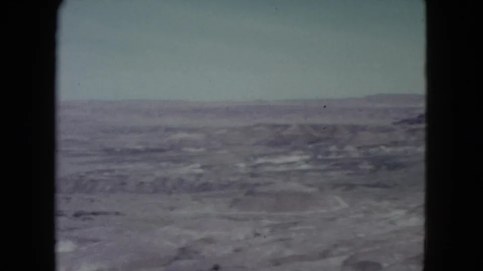 1960 Nevada 8mm frame showing broad rocky desert terrain, a pale sky, and low mountain forms on the horizon.