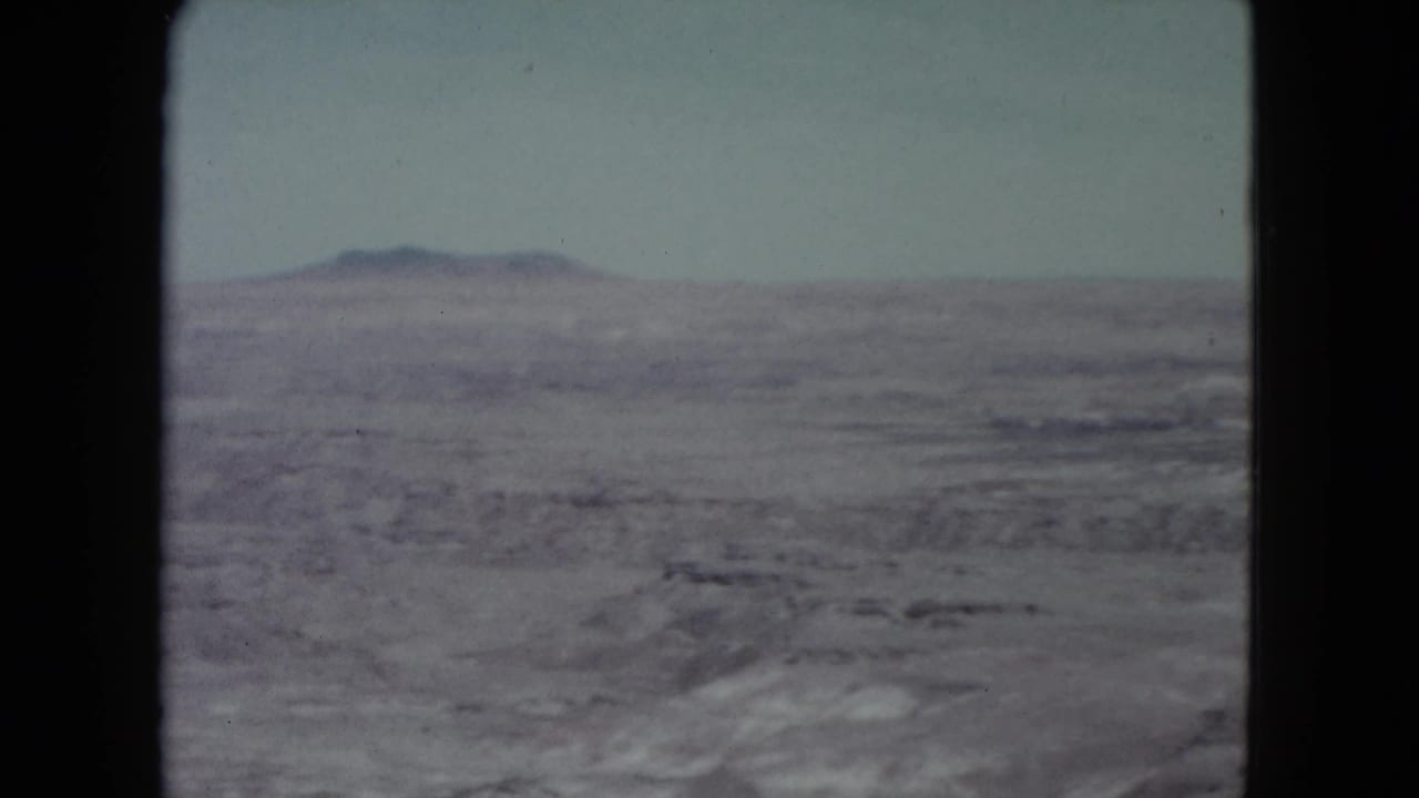 1960 Nevada 8mm view across an open desert basin, with muted rocky ground and a mountain ridge in the distance.