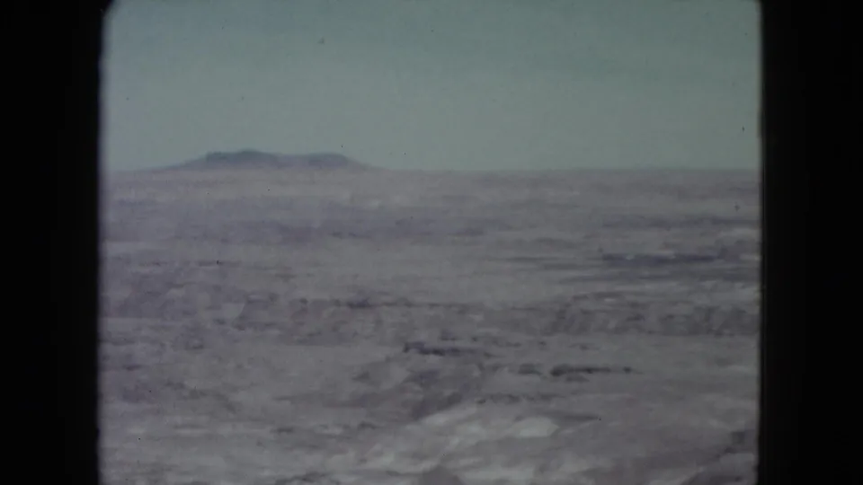 1960 Nevada 8mm view across an open desert basin, with muted rocky ground and a mountain ridge in the distance.