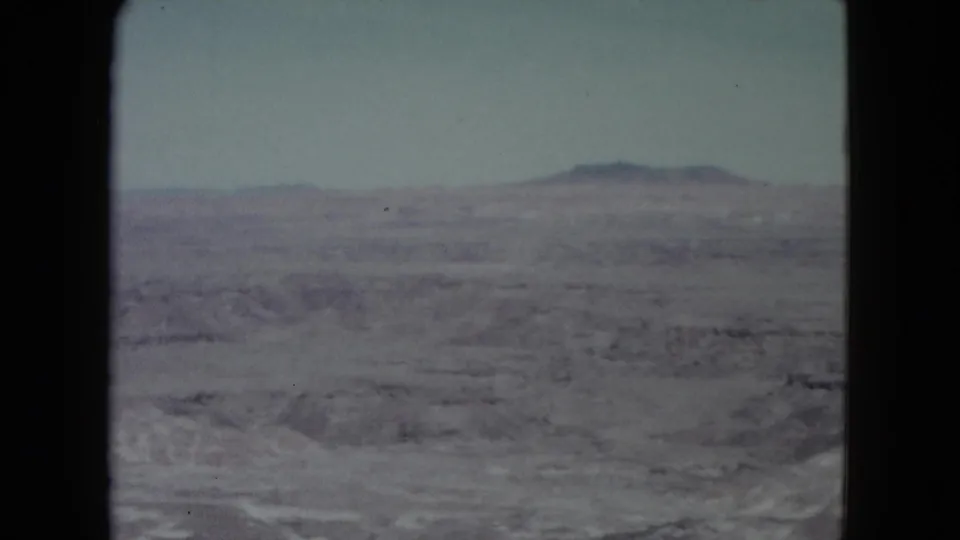 1960 Nevada home-movie frame with hazy desert foreground, low ridges, and distant hills under a washed pale sky.