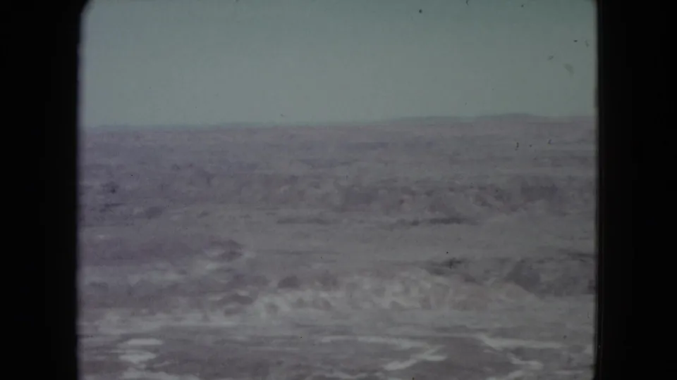 1960 Nevada 8mm landscape showing a rocky desert plain, pale eroded washes, and low mountains along the horizon.