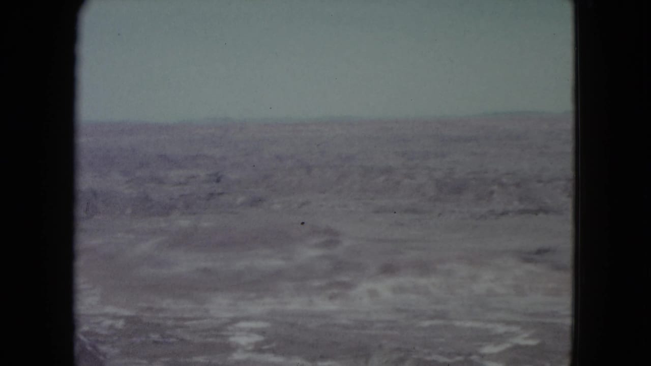 1960 8mm view of open Nevada desert terrain, distant low ridges, and a washed-out sky framed by black film edges.