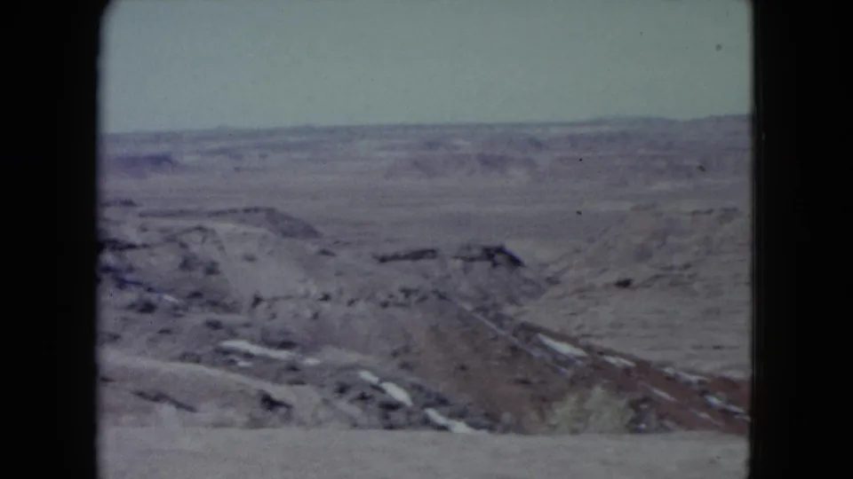 Wide 1960 Nevada landscape frame showing rocky dry ground, desert flats, and far ridges under a washed-out sky.