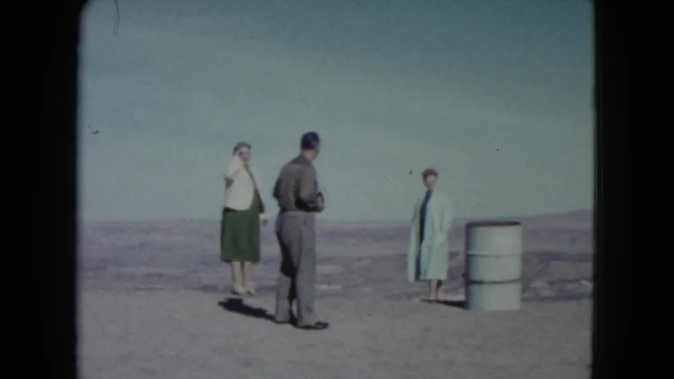 Three adults in 1960 Nevada desert footage stand on open ground near a white barrel under a pale sky.