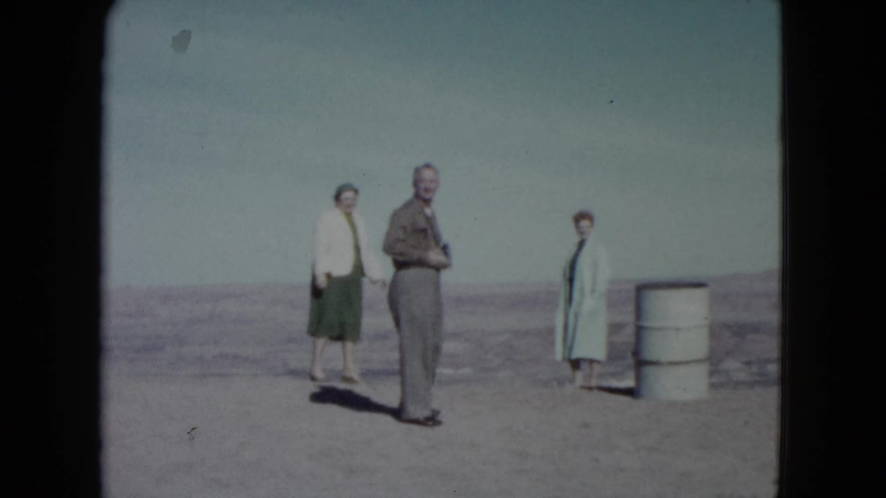 In 1960 Nevada, a man stands between two women on bare desert ground, with a white barrel to the right.