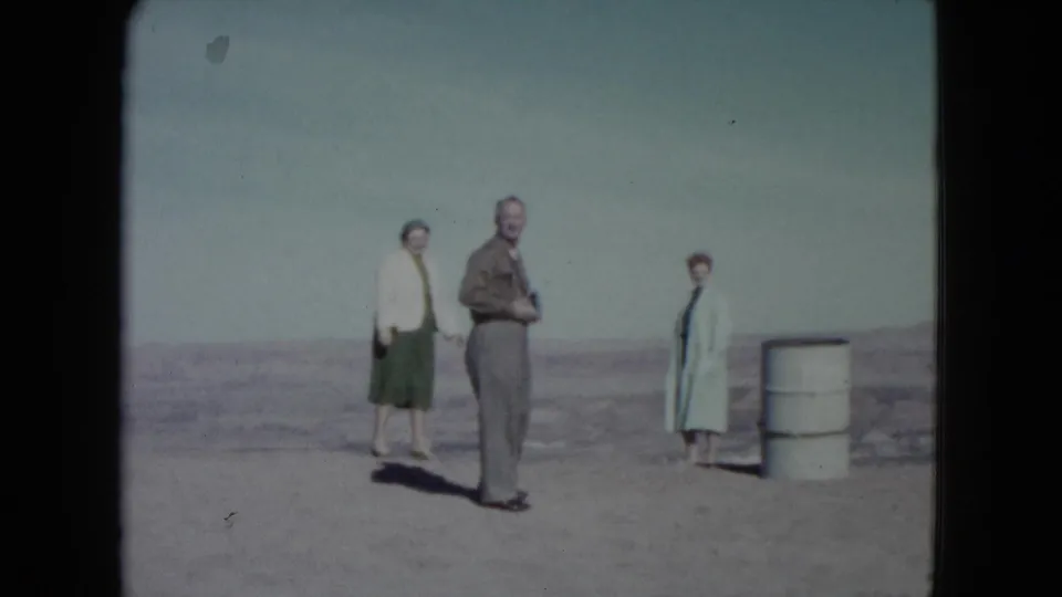 In 1960 Nevada, a man stands between two women on bare desert ground, with a white barrel to the right.