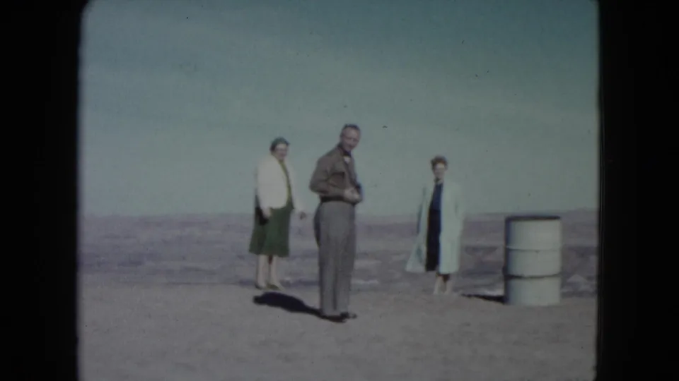A 1960 Nevada home-movie frame shows three adults in desert terrain while the central man looks down at an object.