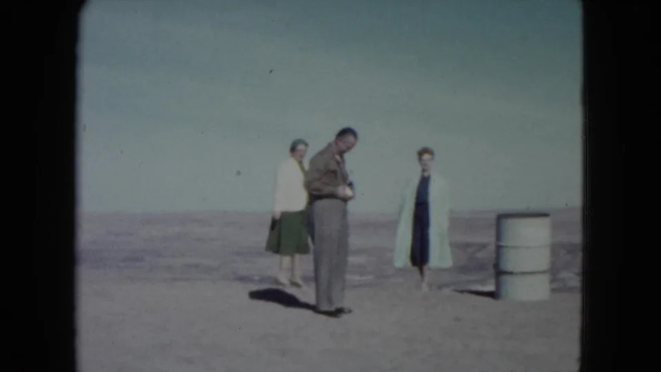 The 1960 Nevada desert scene continues with three adults near a white barrel and a broad, hazy horizon.