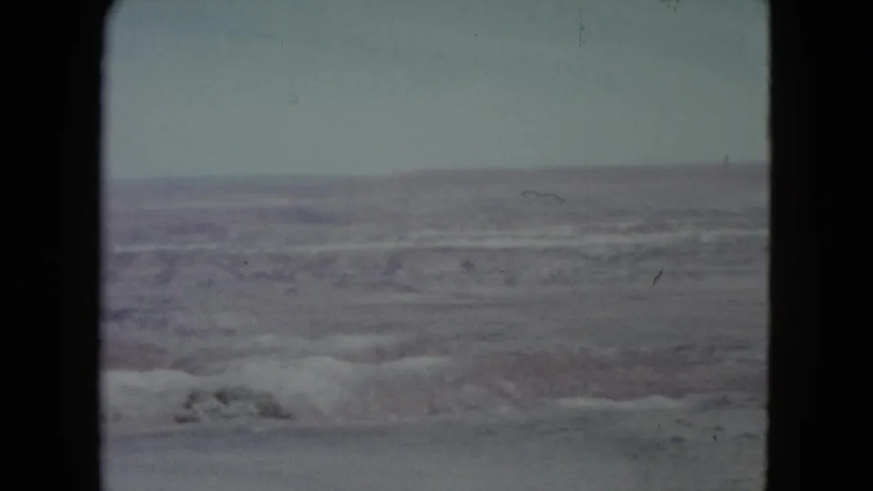 1960 Nevada home-movie view of layered desert canyon terrain beneath a pale sky, bordered by dark film gate edges.