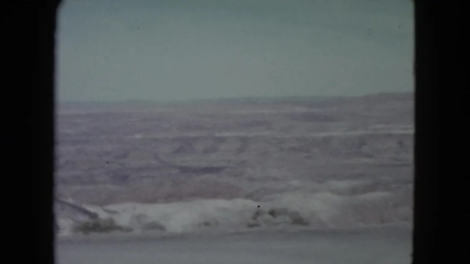 Static 8mm view of a Nevada desert canyon, showing horizontal rock layers, haze, and pale blue sky.
