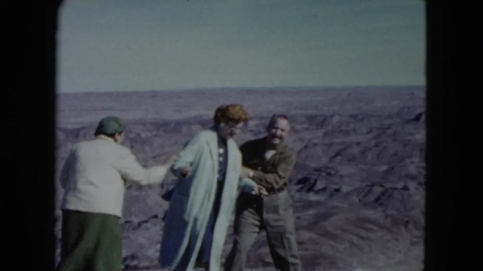 Three adults in period outerwear stand playfully at a Nevada canyon overlook, with desert ridges behind them.