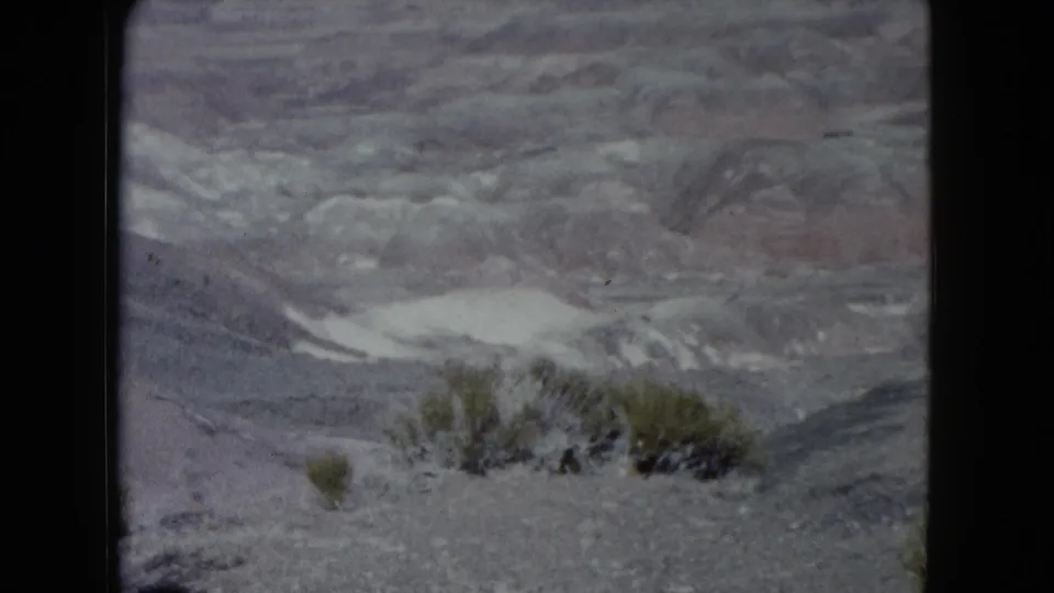 1960 Nevada home-movie frame showing scrub in the rocky foreground and pale desert washes beyond.
