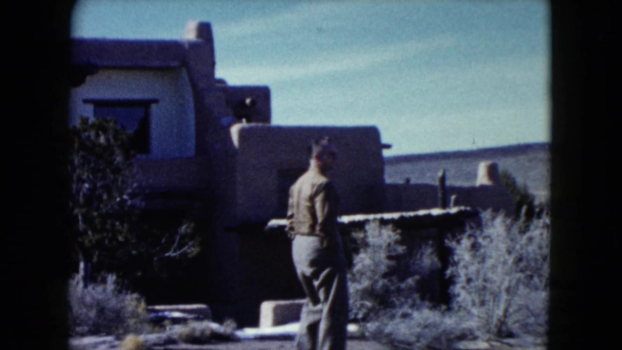 1960 Nevada 8mm frame showing an adult man near earth-toned adobe-style walls, dark windows, vigas, and desert shrubs.