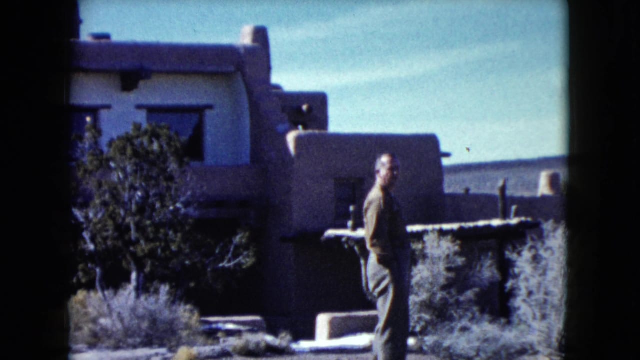 1960 Nevada home-movie frame with an adult man beside an adobe-style building, low wall, dark windows, and sparse desert vegetation.