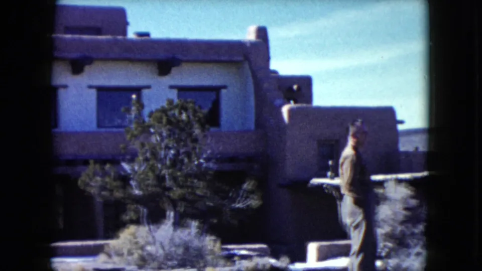 1960 Nevada 8mm frame showing a man moving across the foreground of an adobe-style building with shrubs and shaded window openings.