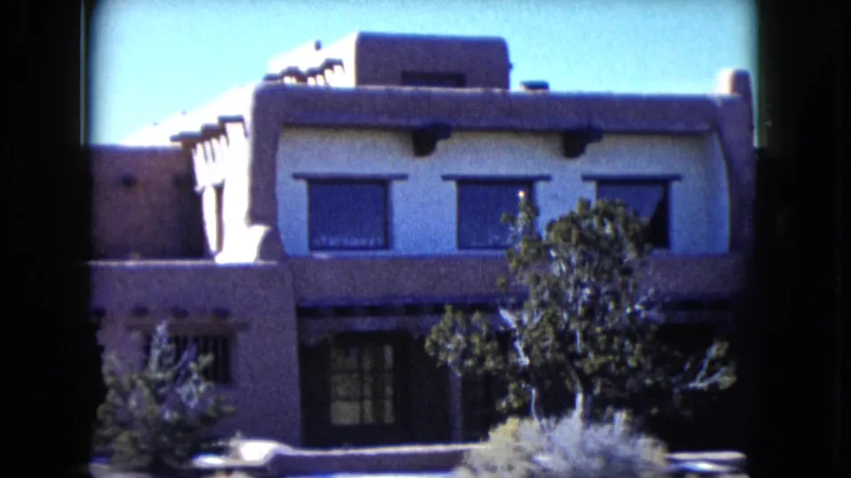 1960 Nevada frame focused on the adobe-style building facade, flat roof, exposed vigas, dark windows, and desert shrubs.