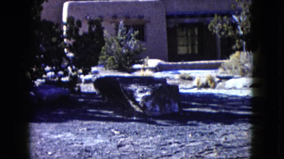 1960 Nevada home-movie frame showing the lower facade of a desert home with shrubs and dry ground in front.
