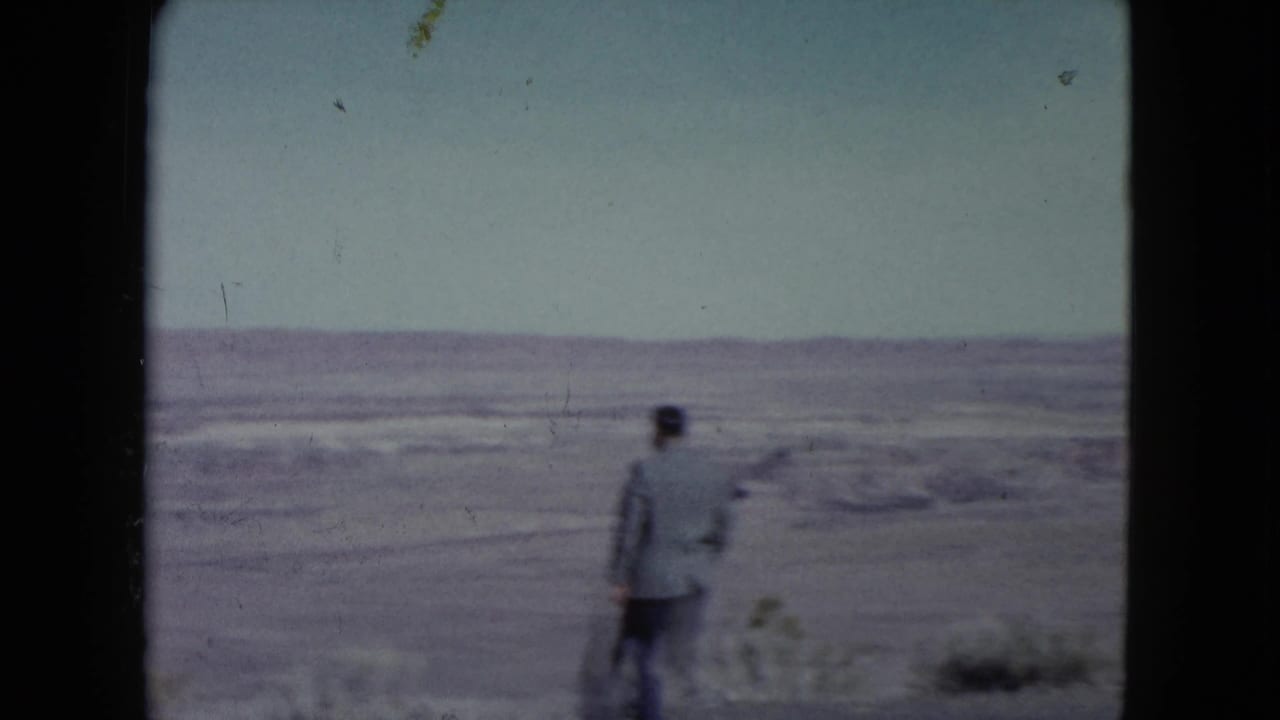 Man in a suit walks away across a sparse desert landscape with distant hills.