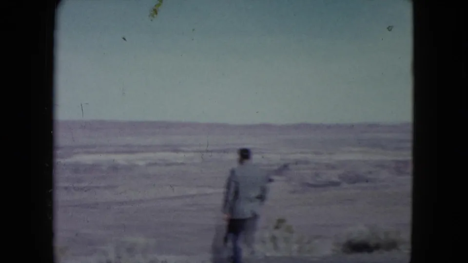 In 1960 Nevada 8mm footage, a suited man walks away from the camera across open desert terrain under a pale sky.